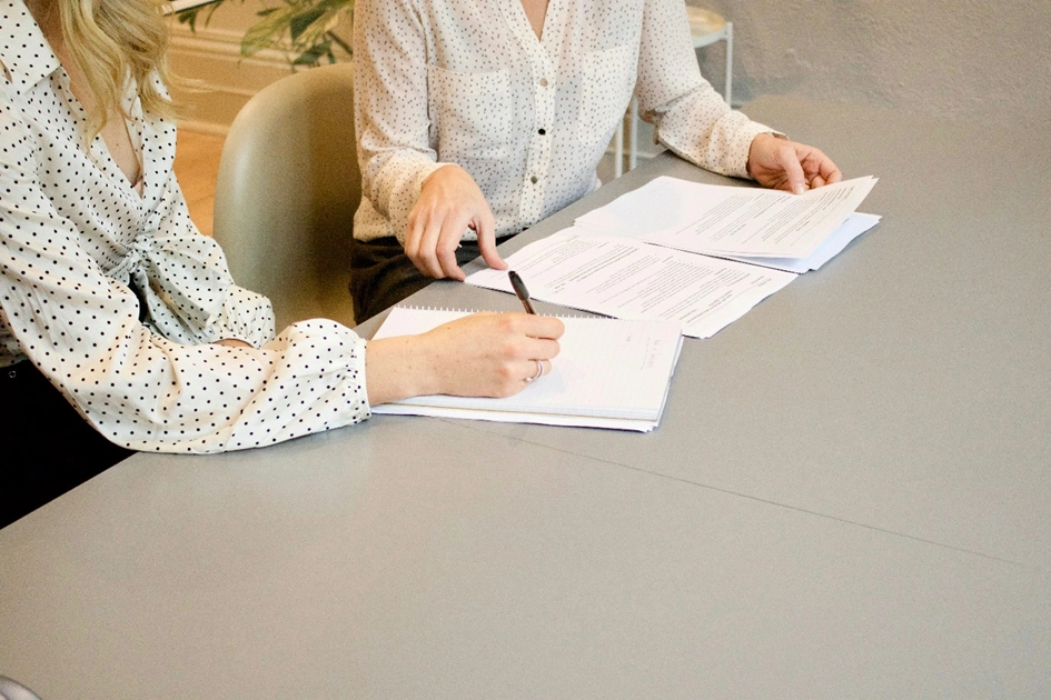 Zwei Frauen sitzen an einem grauen Tisch nebeneinander. Die Gesichter sind nicht zu sehen. Sie gehen einen analogen Arbeitsvertrag durch anstatt einen Digitalen Arbeitsvertrag.