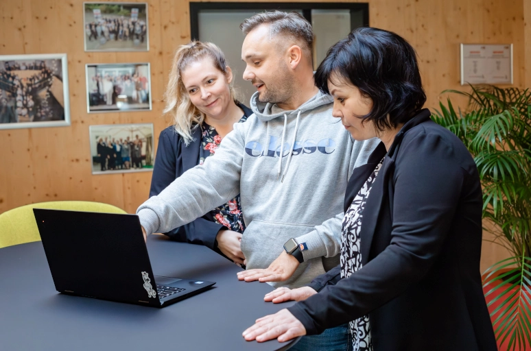 Ein Mann in einem grauen Hoodie steht an einem schwarzen Hochtisch. Neben ihm stehen zwei Kolleginnen in schwarzen Blazern. Vor ihnen steht ein schwarzer Laptop. Der Mann erklärt das Job Sharing.