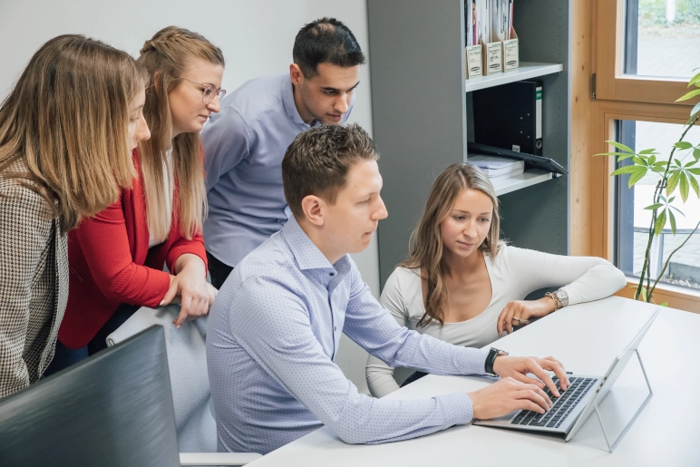 Fünf Mitarbeiter, zwei Männer und drei Frauen, sitzen an einem weißen Tisch. Der Mann im Vordergrund bedient den Laptop. Er berechnet seine Bonuszahlung.