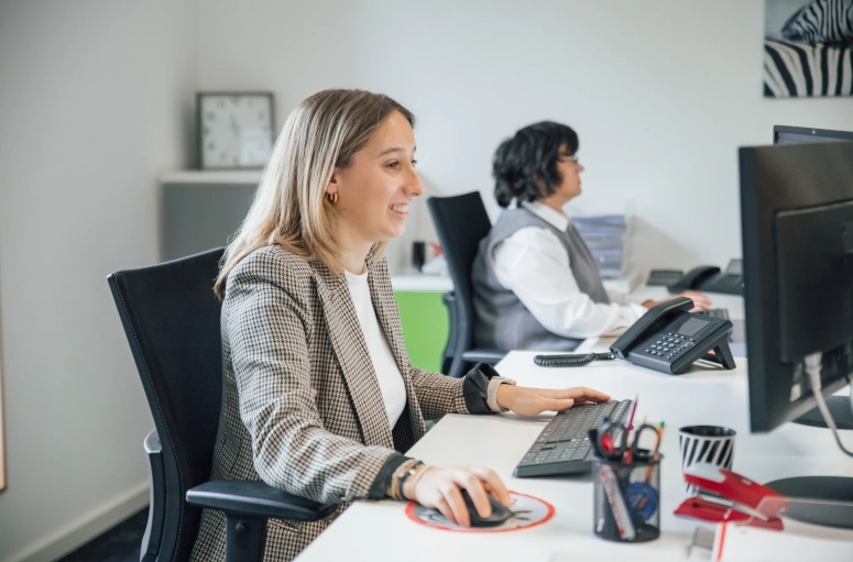 Eine Frau sitzt vor dem PC. Im Hintergrund sieht man eine weitere Frau vor dem PC. Die Frau hält die Maus in der Hand und schaut sich das Thema Arbeitsvertrag Minijob an.