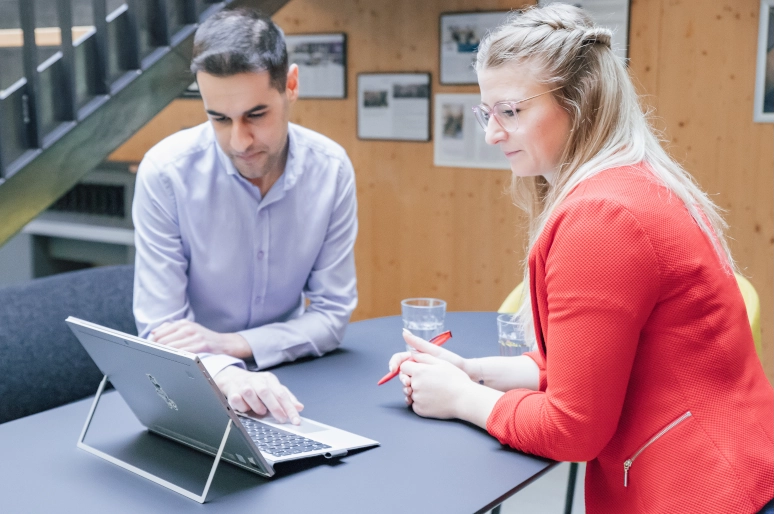 Eine Frau in einem roten Blazer steht an einem schwarzen Hochtisch. Ihr gegenüber steht ein Mann in einem hellblauem Hemd. Sie schauen beide auf einen Laptop und informieren sich über den Zeigarnik-Effekt.