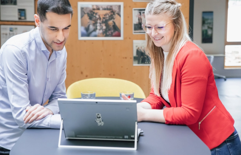 Ein Mann in hellblauem Hemd steht an einem schwarzen Stehtisch neben einer Frau in rotem Blazer. In der Mitte steht ein Laptop. Thema der Diskussion ist Hybrides Arbeiten