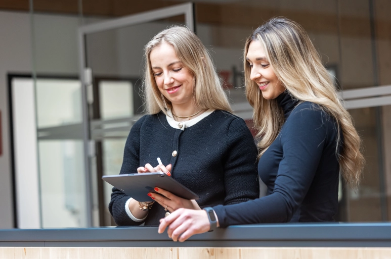 Zwei schwarzgekleidete Frauen stehen vor einem gläserenen Büro. Die Frau links hält ein iPad in der Hand. Beide Frauen nutzen den Arbeitszeitrechner.