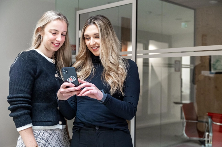 Zwei Frauen in schwarzem Oberteil stehen vor einem gläsernen Büro und schauen auf ein Smartphone. Digitale Lohnzettel sind darauf zu sehen.