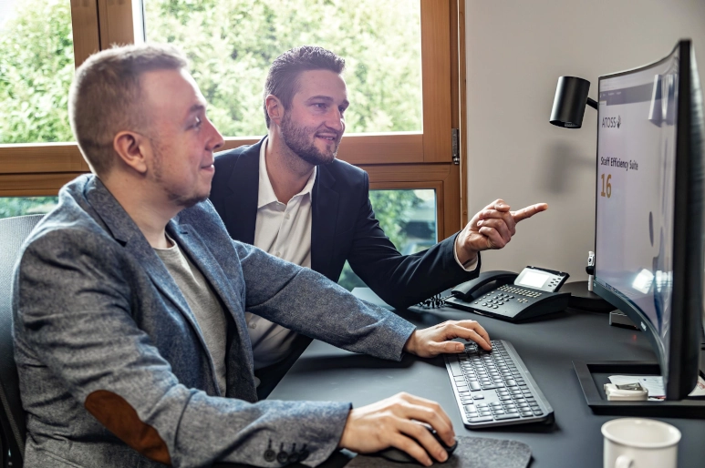 Ein Mann zeigt einem anderen Mann die Software für das Fehlzeitenmanagement auf einem großen Monitor. Beide sitzen vor einem schwarzen Schreibtisch.