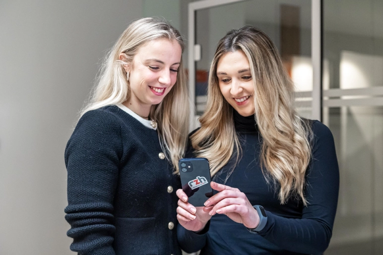 Zwei Frauen in schwarz gekleidet, stehen vor einem gläsernen Büro und schauen beide auf das iPhone der rechten Frau. Sie sprechen über Teilzeit in Elternzeit.