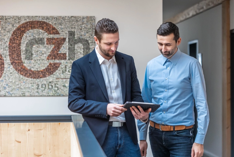 Ein Mann in einem schwarzen Blazer zeigt einem anderen Mann in hellblauem Hemd digitale Schichtmodelle auf seinem iPad.