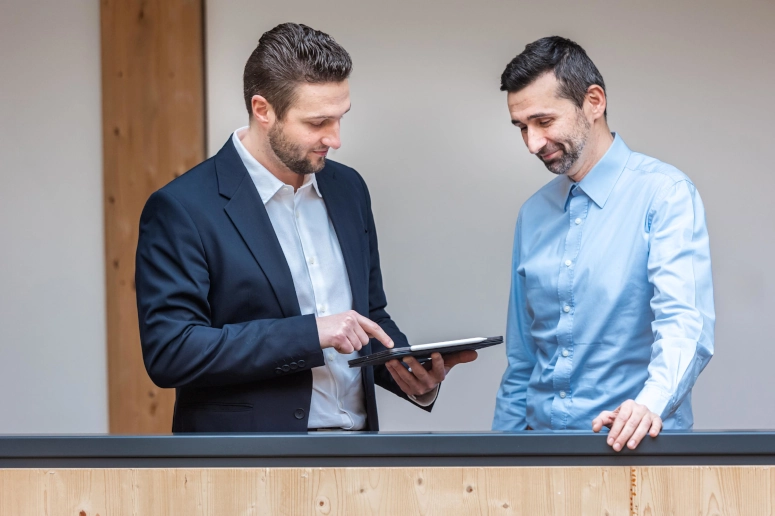 Zwei Experten für Arbeitszeitmodelle diskutieren an einer Balustrade über das Thema. Der Mann links hat einen schwarzen Blazer an, der Mann rechts ein hellblaues Hemd. Der Mann links hält zudem ein iPad in der Hand.