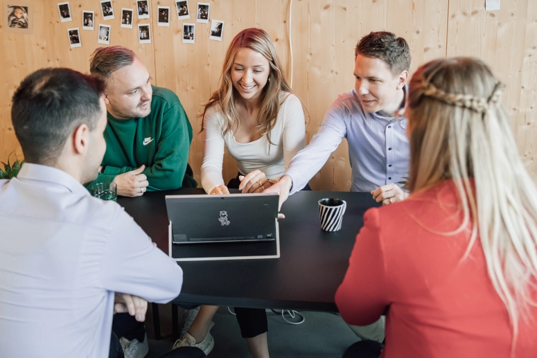 Eine Gruppe von Menschen (drei Männer und zwei Frauen) sitzen an einem Tischen und besprechen das Thema Kurzarbeit. In der Mitte steht ein Laptop, auf den ein Mann und eine Frau zeigen.