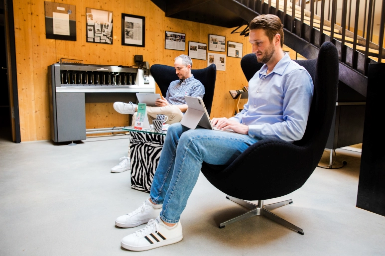 Zwei Männer in hellblauem Hemd sitzen jeweils in einem schwarzen Sessel. Sie haben beide einen Laptop in der Hand, der Teil eines Incentives Programm war. Im Hintergrund ist eine Holzwand zu sehen, an der verschiedene Bilder hängen.