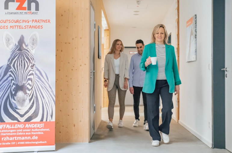 Zwei Frauen und ein Mann gehen einen Flur entlang. Die Frau im Vordergrund trägt eine Tasse. Sie unterhalten sich über Job Shadowing.