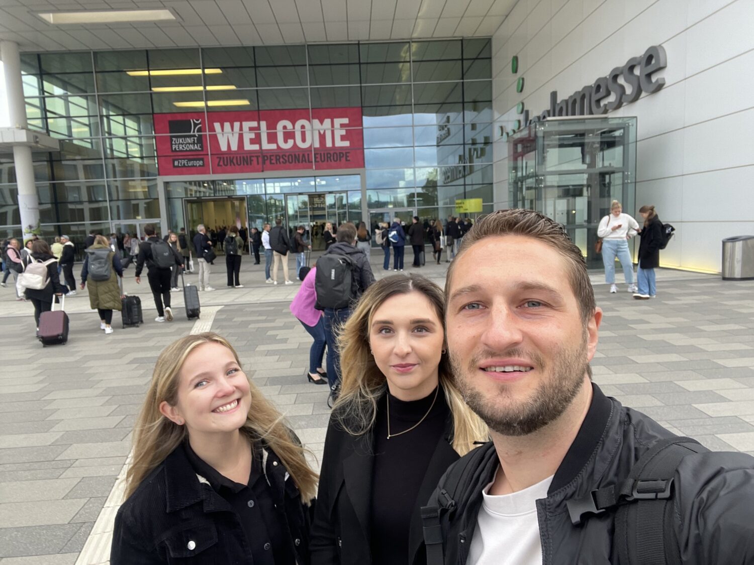 drei Mitarbeiter, die vor der Messe Zukunft Personal stehen und ein Selfie machen