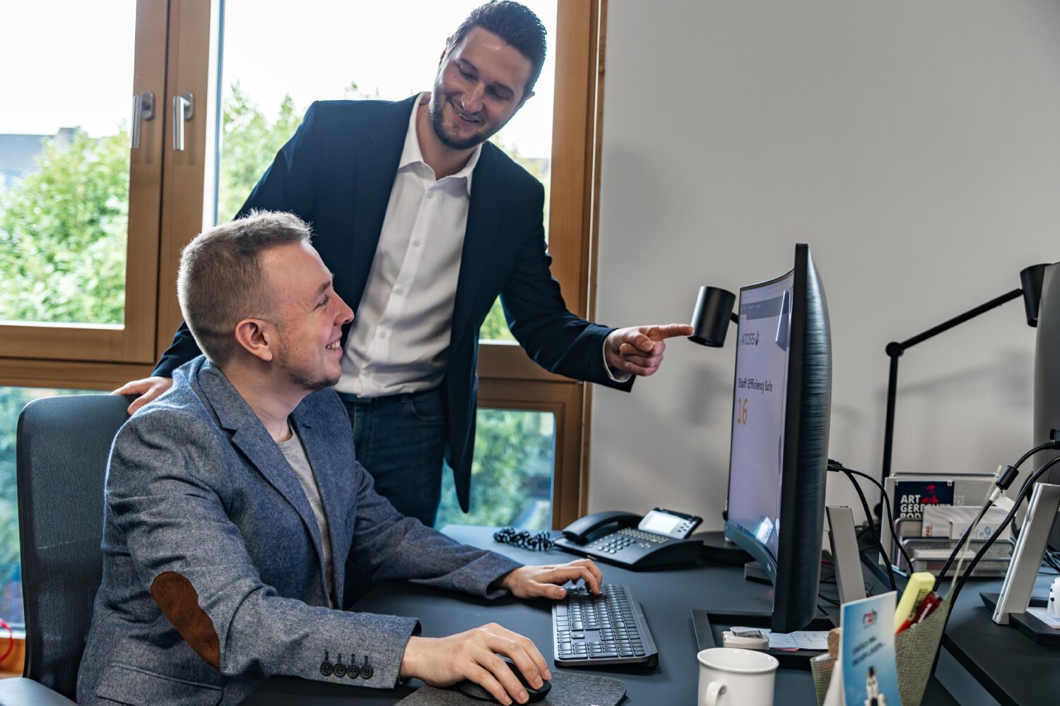 Zwei Arbeitskollegen sitzen im Büro vor Computer und einer zeigt dem anderen etwas.