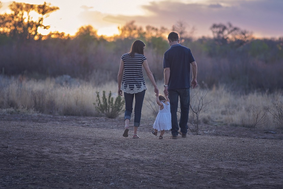 Die Familienstartzeit würde es wie im Bild Vater, Mutter und Kind ermöglichen, während des Sonnenaufgangs auf einem Feld spazieren zu gehen. Im Hintergrund sind Bäume zu sehen.