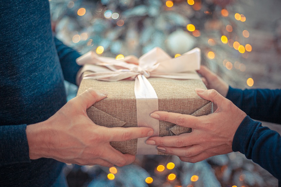 Man sieht zwei Hände, die ein beiges personalisiertes Geschenk an zwei andere Hände übergeben. Im Hintergrund ist ein schneebedeckter Weihnachtsbaum
