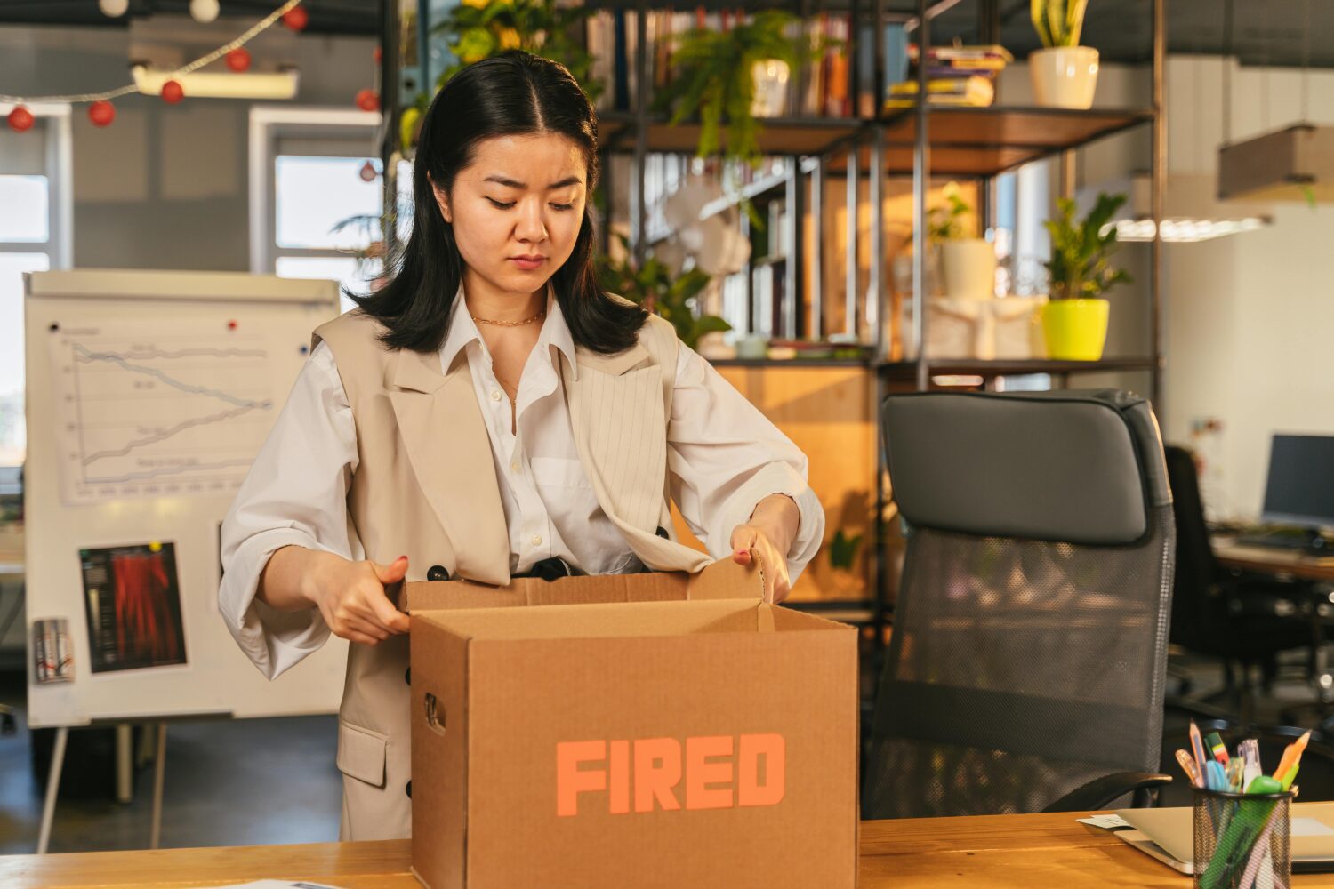 Abgebildet ist eine braunhaarige Frau, die im Büro ihre Sachen in einen Karton packt, auf dem steht "Fired".