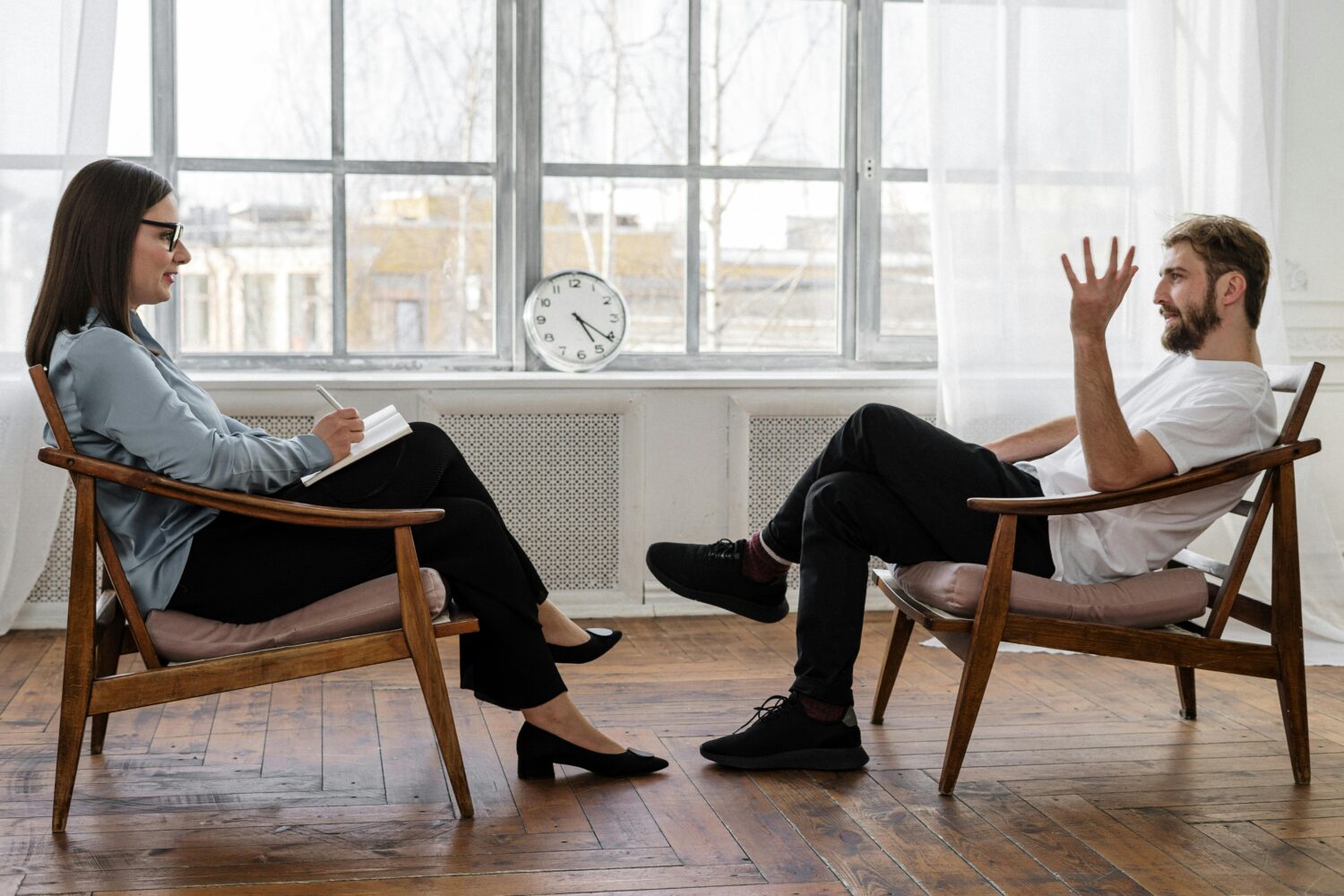 Coaching dargestellt durch eine Frau als Coach und einen Mann als gecoachte Person, die sich auf zwei Stühlen gegenüber sitzen.