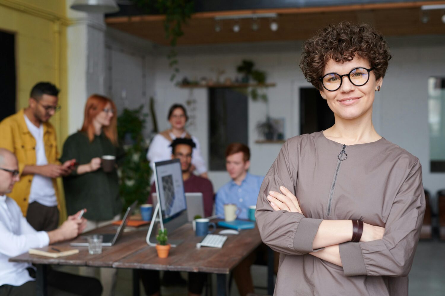 Eine Frau als Führungskraft steht im Vordergrund, im Hintergrund sind Mitarbeitende an einem Tisch mit Laptops bei der Arbeit zu sehen.