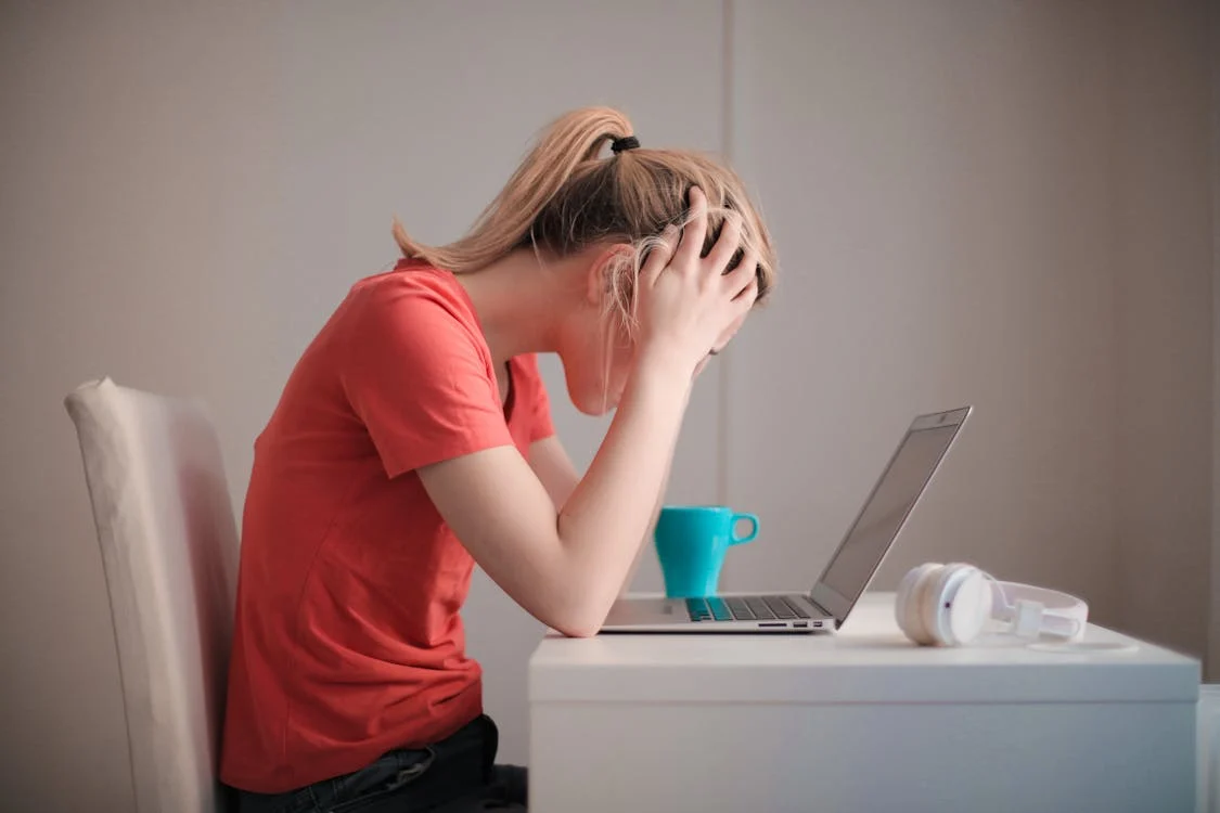 Eine Frau in orangenem Oberteil sitzt an einem weißen Tisch und schaut auf den Laptop. Sie ist verzweifelt wegen Gewalt gegen Frauen