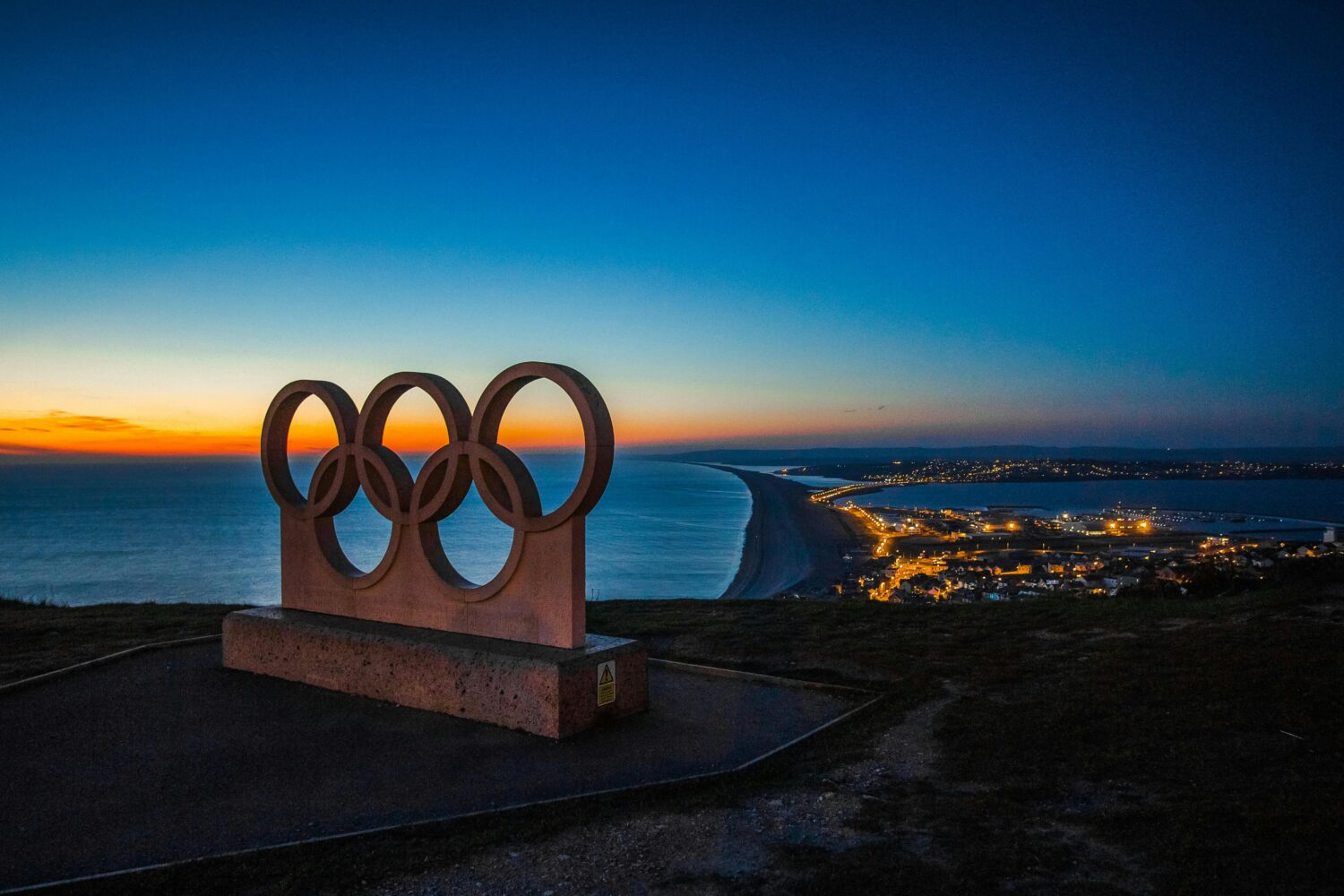 Olympische Ringe als Statue auf einem Hügel, im Hintergrund Meer mit einer Stadt und Sonnenuntergang