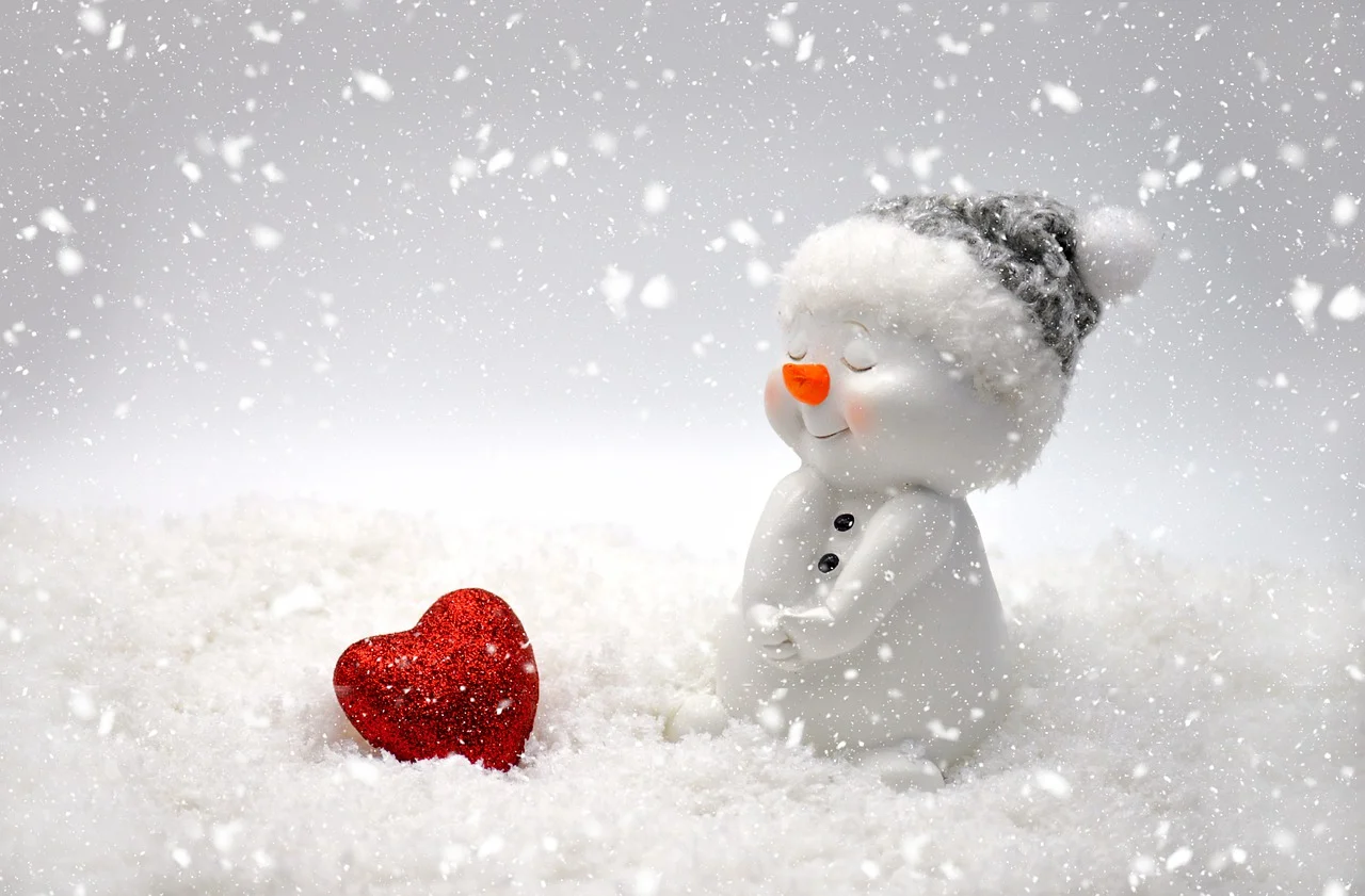 Ein kleiner Schneemann hat eine Mütze auf, sitzt im Schnee und hat ein rotes Herz direkt vor sich. Das Bild symbolisiert Weihnachten.